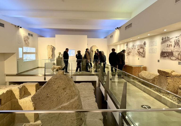 Interior view of a modern museum in Spain with a raised glass walkway. Hartwick students and faculty look at ancient stone sculptures and wall illustrations. Large stone blocks are visible on the lower level.