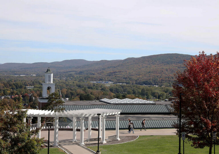 Elevated view of Hartwick Colleg campus with a white pergola, a clock tower, and students walking, set against a backdrop of autumn-colored mountains under a blue sky.