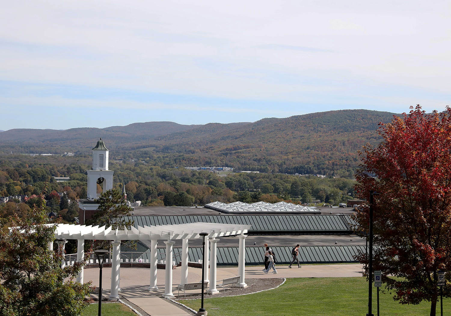 Elevated view of Hartwick Colleg campus with a white pergola, a clock tower, and students walking, set against a backdrop of autumn-colored mountains under a blue sky.