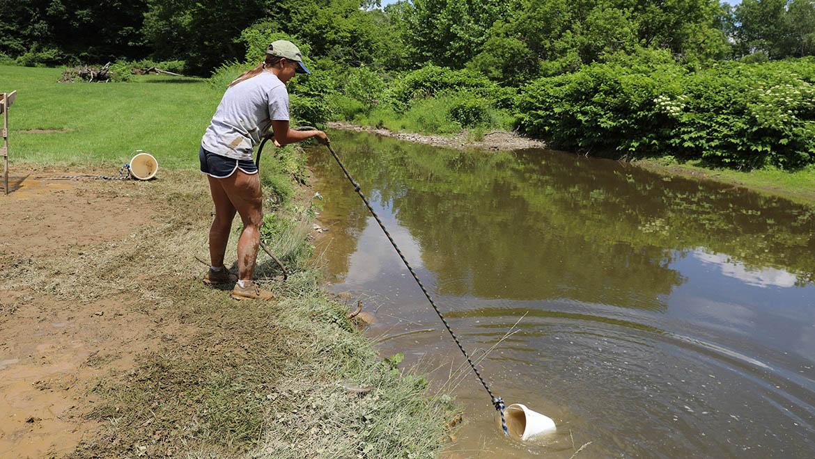 Field Archaeology student gathering water in bucket at excavation site at Hartwick College's Pine Lake Environmental Campus dig