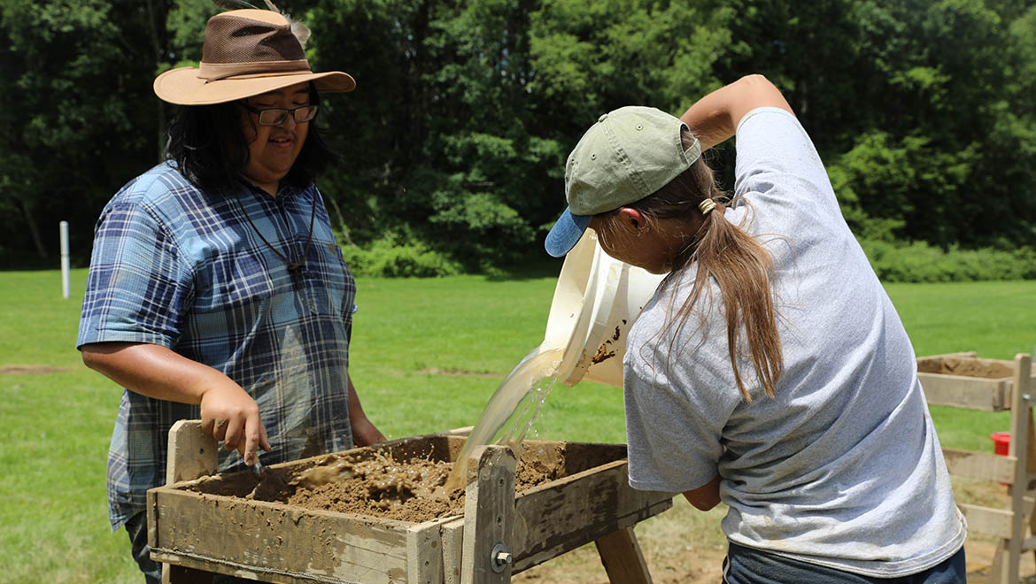 Field Archaeology student poring water at excavation site at Hartwick College's Pine Lake Environmental Campus dig to fins artifacts in soil