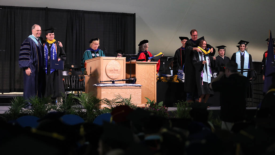 Hartwick College graduates walking to the stage to receive their hood and diploma and to be congratulated by Interim President Mullen.
