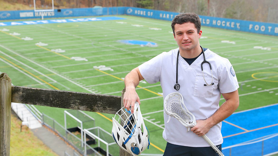 Colby Simonetti '26 with lacrosse stick, helmet wearing scrubs with Hartwick College's Wright Stadium in background