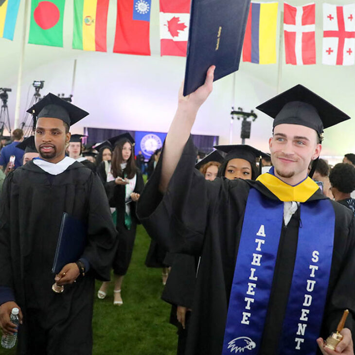 Hartwick College graduates of the Class of 2025 holding up diploma and alumni bell marching out of tent after Commencement ceremony