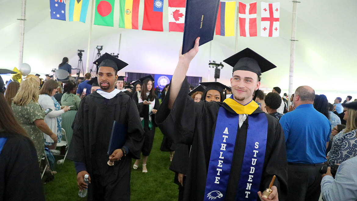 Hartwick College graduates of the Class of 2025 holding up diploma and alumni bell marching out of tent after Commencement ceremony