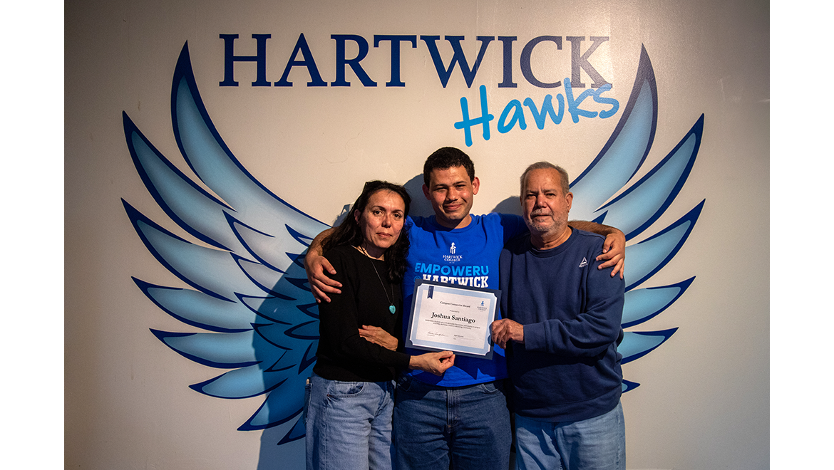 EmpowerU student with family for photo with Hartwick Hawks Wings mural