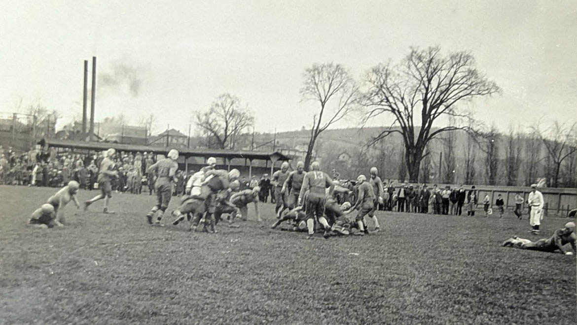 Black and white photo of an old-fashioned American football game with players in a scrum, spectators in grandstands, and industrial smokestacks in the background.