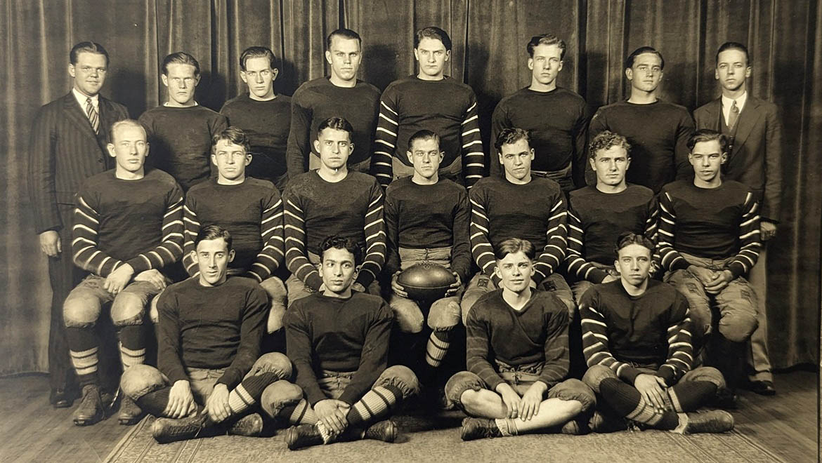 A black and white photo of nineteen young men, a vintage football team, in three rows. Most wear dark, striped long-sleeved jerseys and padded pants. Two men in the back wear suits, and one player holds a football.