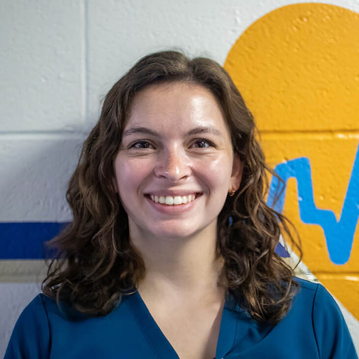 Portrait of Emilyann Feldman standing in front of a yellow heart mural with a blue ECG line.