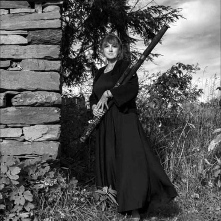 Black and white photo of a Pamela WEst-Finkle holding a long, dark woodwind instrument outdoors, standing next to a stone wall.