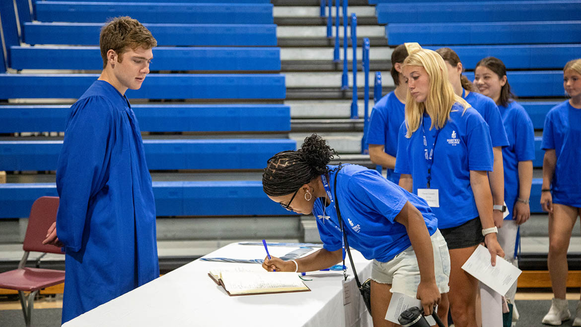 Hartwick student in a blue graduation gown stands near a white table where a young woman in a blue t-shirt signs the Hartwick College Matriculation book. Other students in blue t-shirts stand in the background, with blue bleachers behind them.