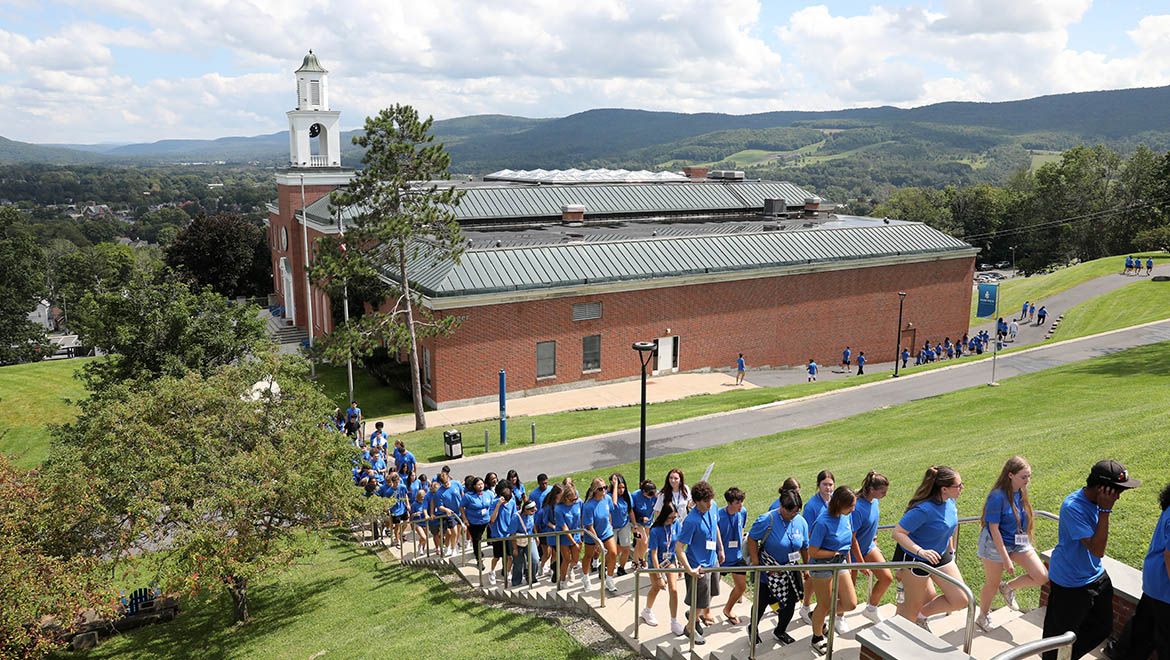 New Hartwick College students in blue shirts walks up an outdoor staircase on a campus, with a brick building, clock tower, and distant hills visible.
