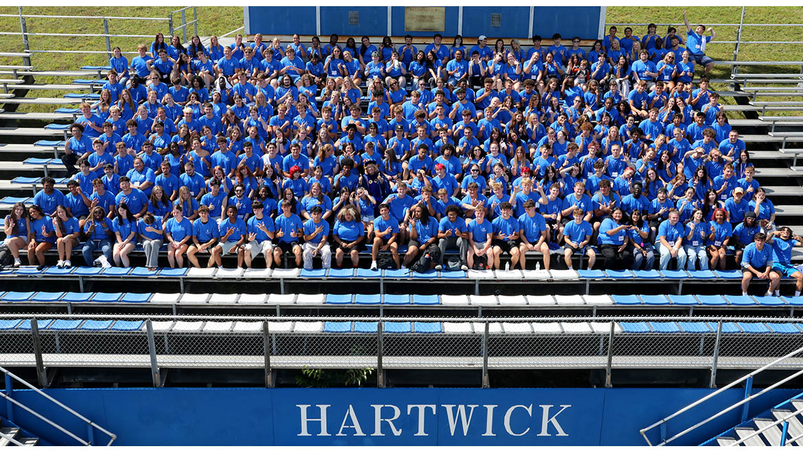 New Hartwick College students wearing blue t-shirts seated on blue bleachers, with 'HARTWICK' written on a blue wall below them.