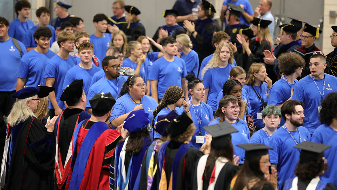 New Hartwick College students in blue t-shirts and adults in academic regalia are gathered indoors, with some faculty clapping.