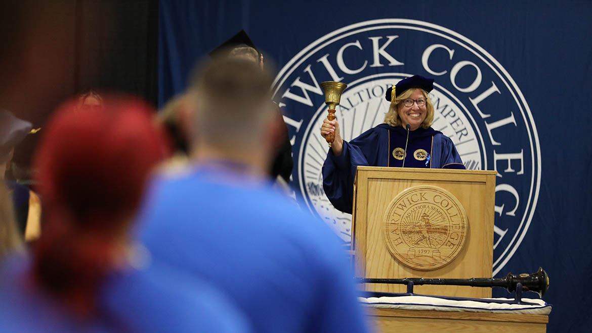Hartwick College President Laurel Bongiorno in academic regalia rings a the Harwick bell behind a podium to being Opening Convocation ceremony for new students with the Hartwick College seal, against a blue banner.