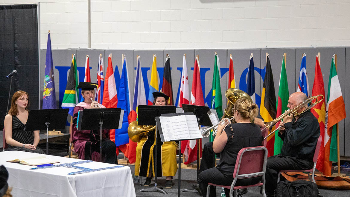 Hartwick College brass ensemble performs during Opening Convocation in front of international flags. Two musicians wear academic gowns, playing trumpet and tuba. Two others play trumpet and trombone. A woman sits to the left of the ensemble, and a table with a book and blue object is in the foreground.