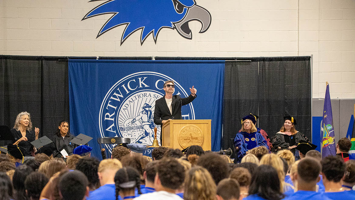 Assistant Professor of Sociology Zachary McKenney speaks at a podium in front of a Hartwick College banner, with an audience in the foreground and people in academic regalia seated on stage during Opening Convocation.