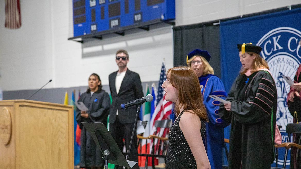 Hartwick student sings Hartwick College alma mater into a microphone at a podium with Opening Convocation platform party and flags and a blue banner are in the background.