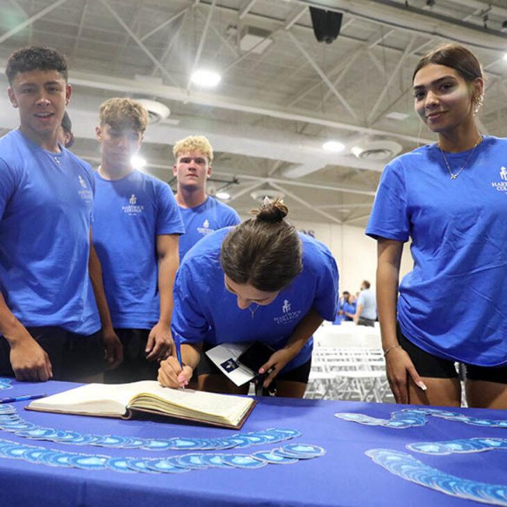 Five students in blue Hartwick College t-shirts at a table, one signing a book.