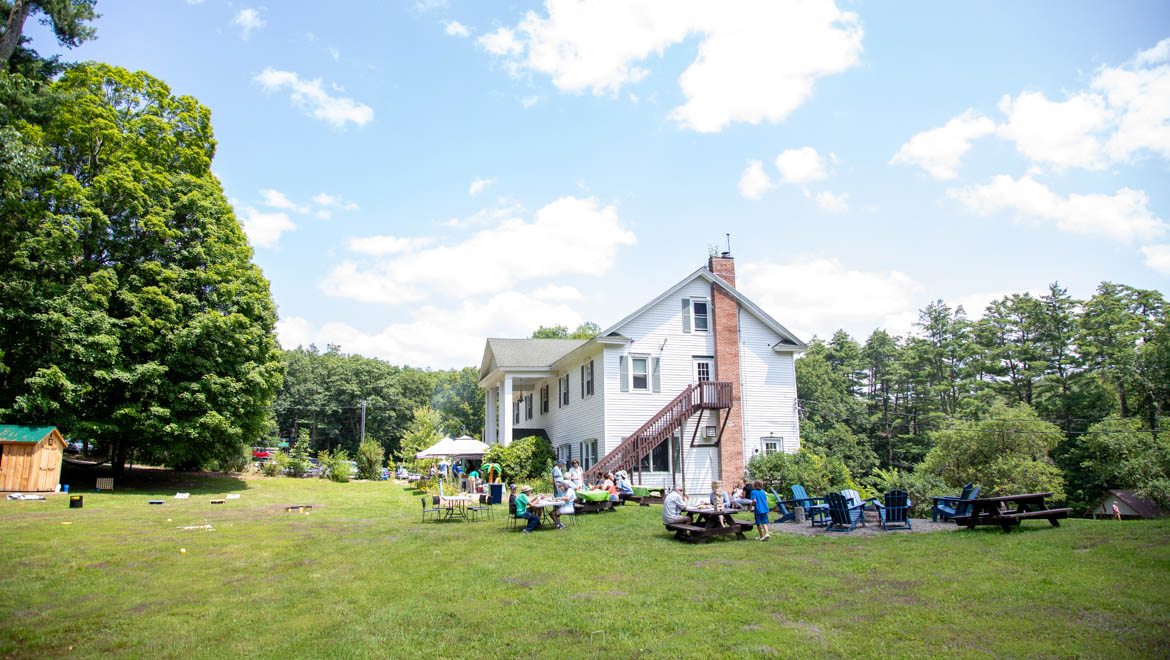 Hartwick College Pine Lake Robertson Lodge surrounded by a green lawn where people are gathered at picnic tables and under a tent on a sunny day.