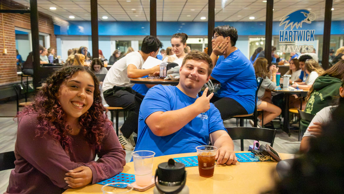 Two smiling Hartwick students, a woman with curly hair and a man in a blue shirt making a peace sign, sit at a table with bingo cards and drinks in a busy common area with a 'Hartwick Hawks' sign in the background.