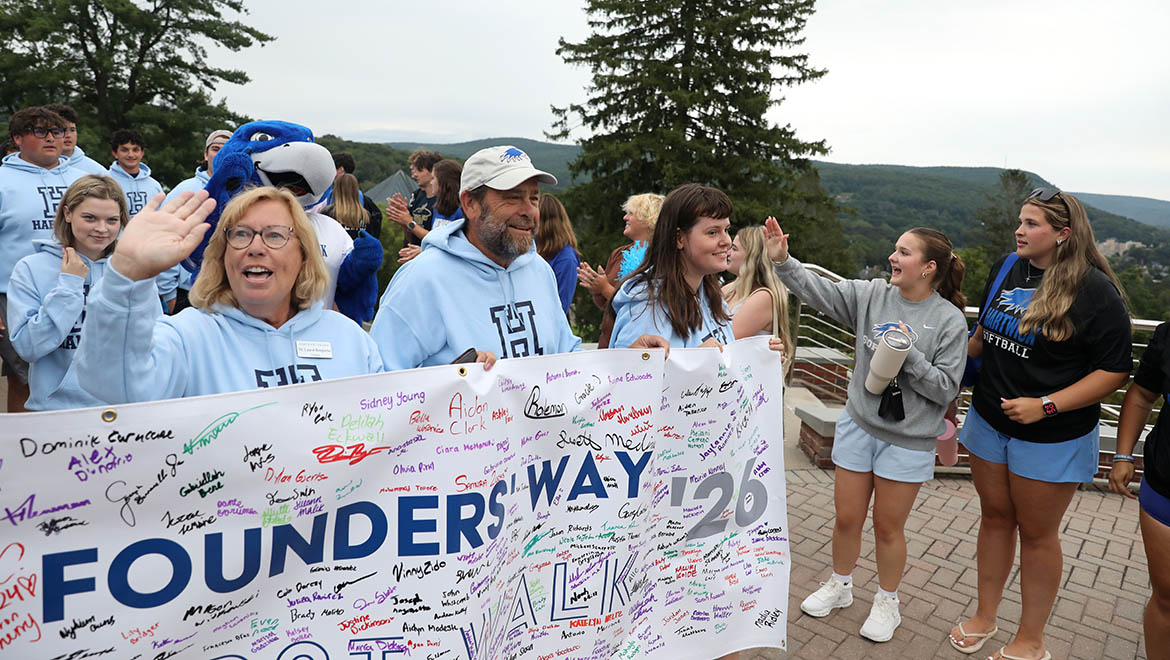 Hartwick new students with President Bongiorno, including Swoop the Hawks mascot, holding a signed banner that reads 'Founders Way Walk '26' outdoors on a college campus with hills in the background. A woman in a light blue hoodie waves to the camera.