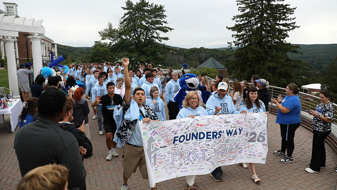 Hartwick College new students many in light blue hoodies, walk on a paved path carrying a banner that reads 'Founders' Way First Walk '26' with signatures. A blue bird mascot- Swoop- is among them.