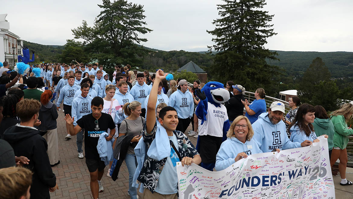 Hartwick College new students many in light blue hoodies, walk on a paved path carrying a banner that reads 'Founders' Way First Walk '26' with signatures. A blue bird mascot- Swoop- is among them.