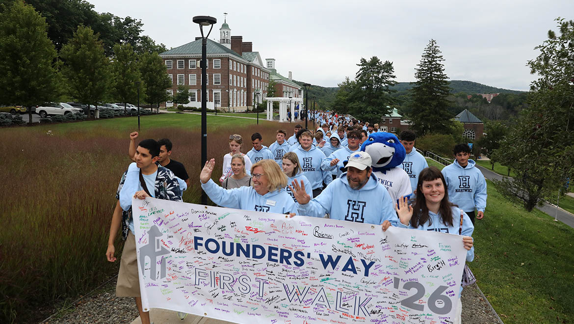 Hartwick College new students many in light blue hoodies, walk on a paved path carrying a banner that reads 'Founders' Way First Walk '26' with signatures. A blue bird mascot- Swoop- is among them.