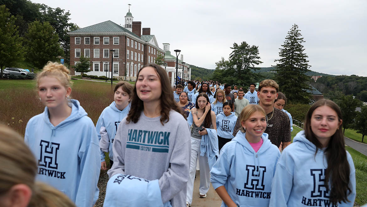 A group of Hartwick students wearing light blue Hartwick hoodies walk on a campus pathway-Founders' Way, with a historic brick building in the background during First Walk