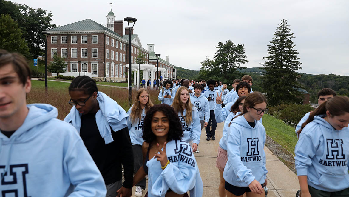 A group of Hartwick students wearing light blue Hartwick hoodies walk on a campus pathway-Founders' Way, with a historic brick building in the background during First Walk