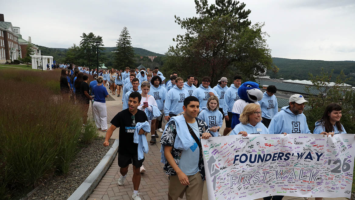 Hartwick College new students many in light blue hoodies, walk on a paved path carrying a banner that reads 'Founders' Way First Walk '26' with signatures. A blue bird mascot- Swoop- is among them.