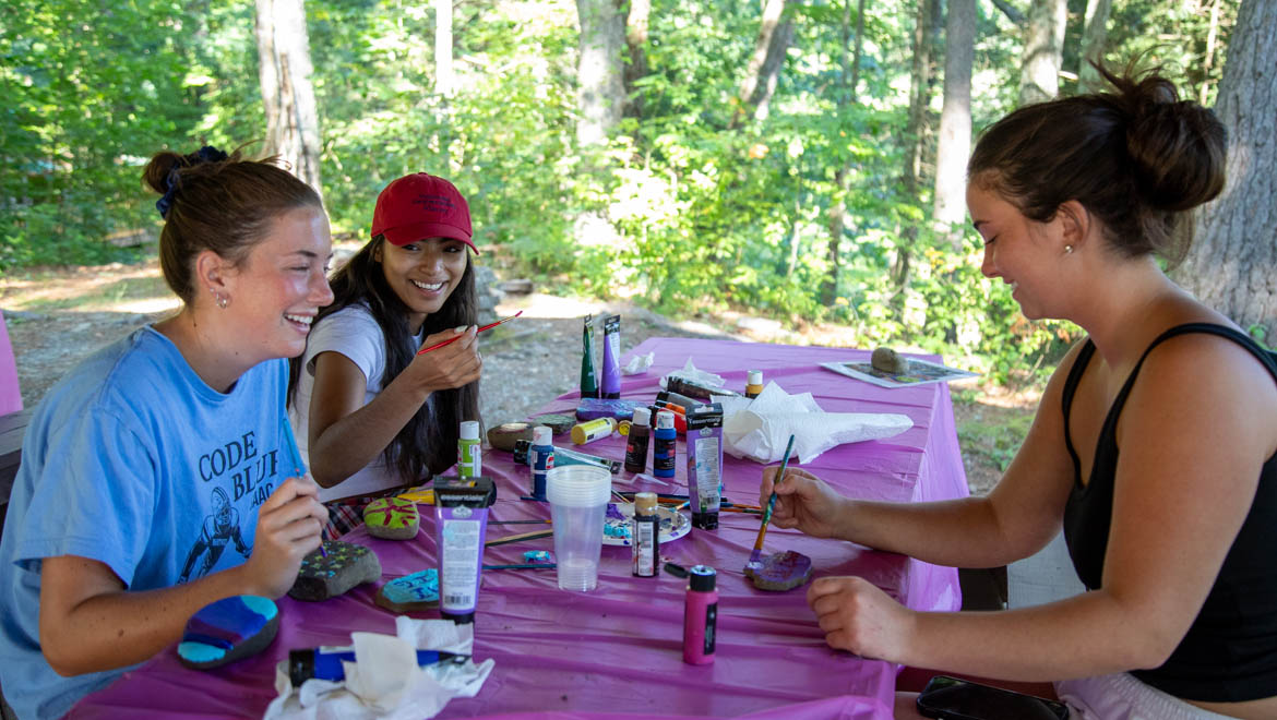 Three young women at Pine Lake smiling and painting rocks at an outdoor table covered with art supplies.