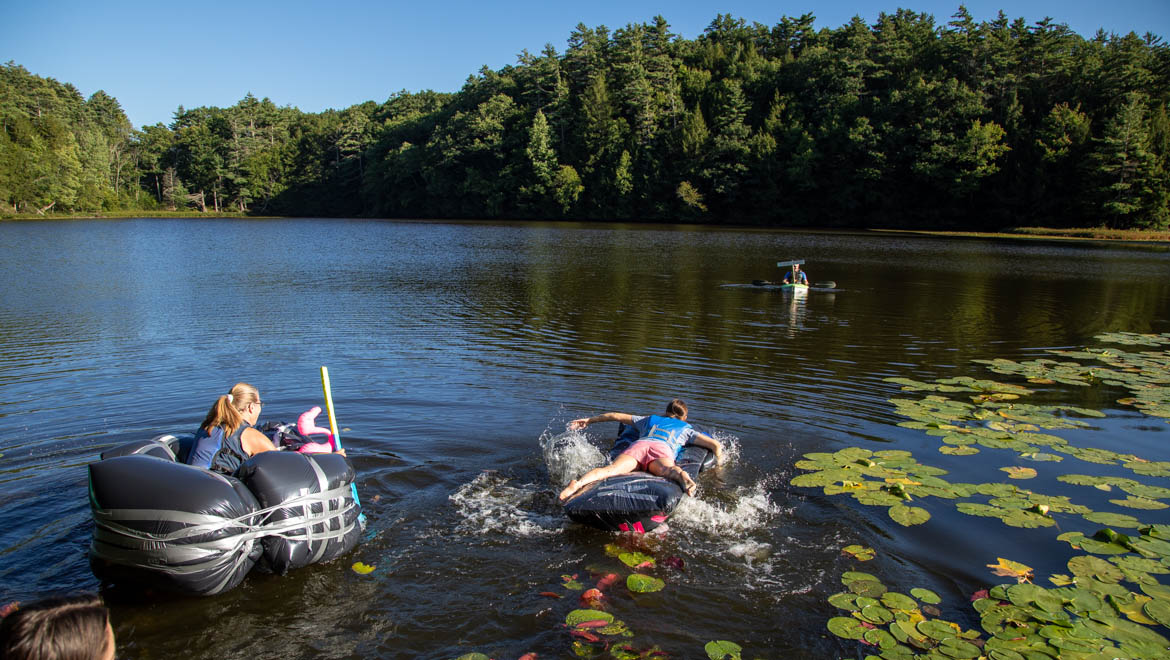Two people on homemade inflatable rafts on a lake, with a third person in a small boat in the distance on Pine Lake. The lake is surrounded by a green forest under a clear sky, with lily pads in the foreground.