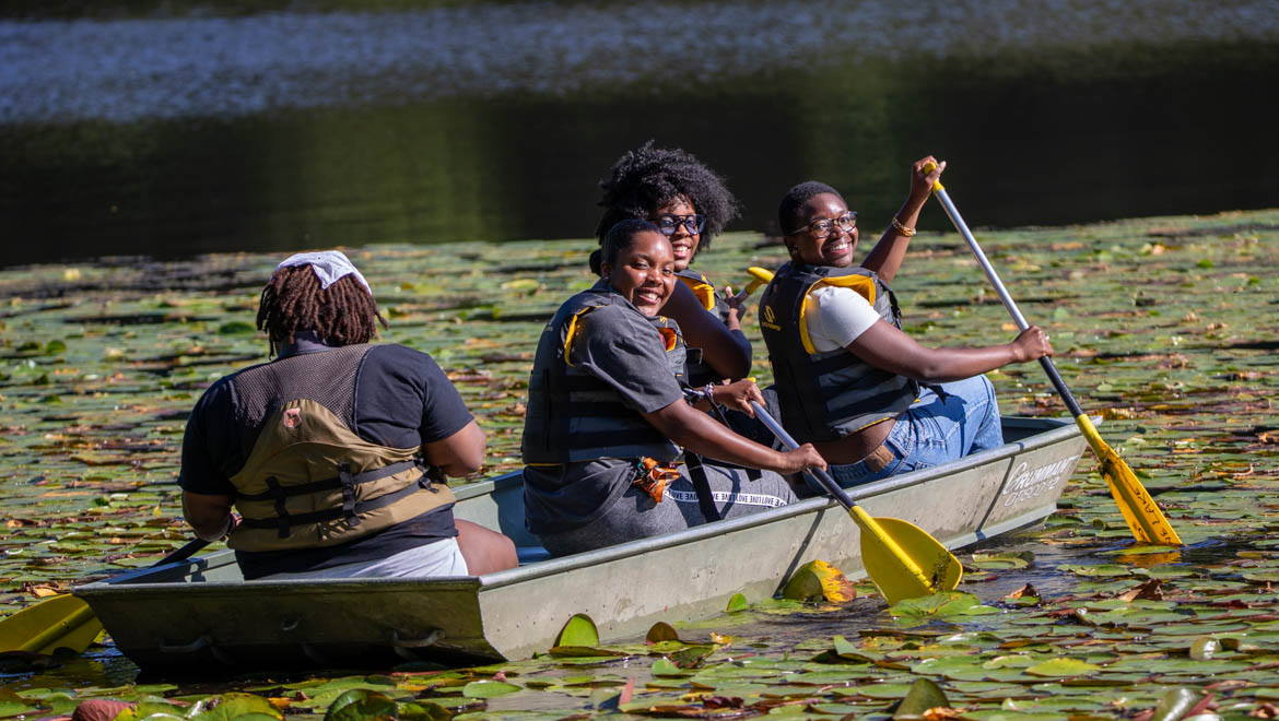 Four Hartwick students in a small boat on a Pine Lake covered with lily pads, wearing life vests and paddling, smiling happily.