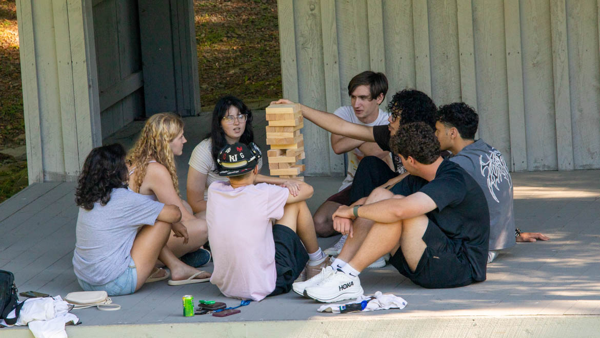 Seven young adults sitting in a circle on a wooden stage at Pine Lake, playing Jenga. One person is reaching for a block from the tall wooden tower.