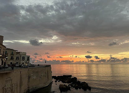 A coastal town in Sicily at sunrise or sunset, with light-colored buildings on a cliff overlooking calm water reflecting an orange and yellow sky with grey clouds.