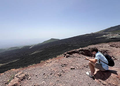 A man sits on rocky ground, looking at his phone, with a vast volcanic landscape and hazy sky in the background.