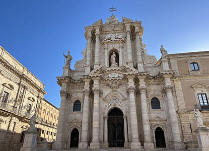 A grand, ornate Baroque church facade made of light stone, featuring multiple columns, statues, and a cross on top, against a clear blue sky in Sicily