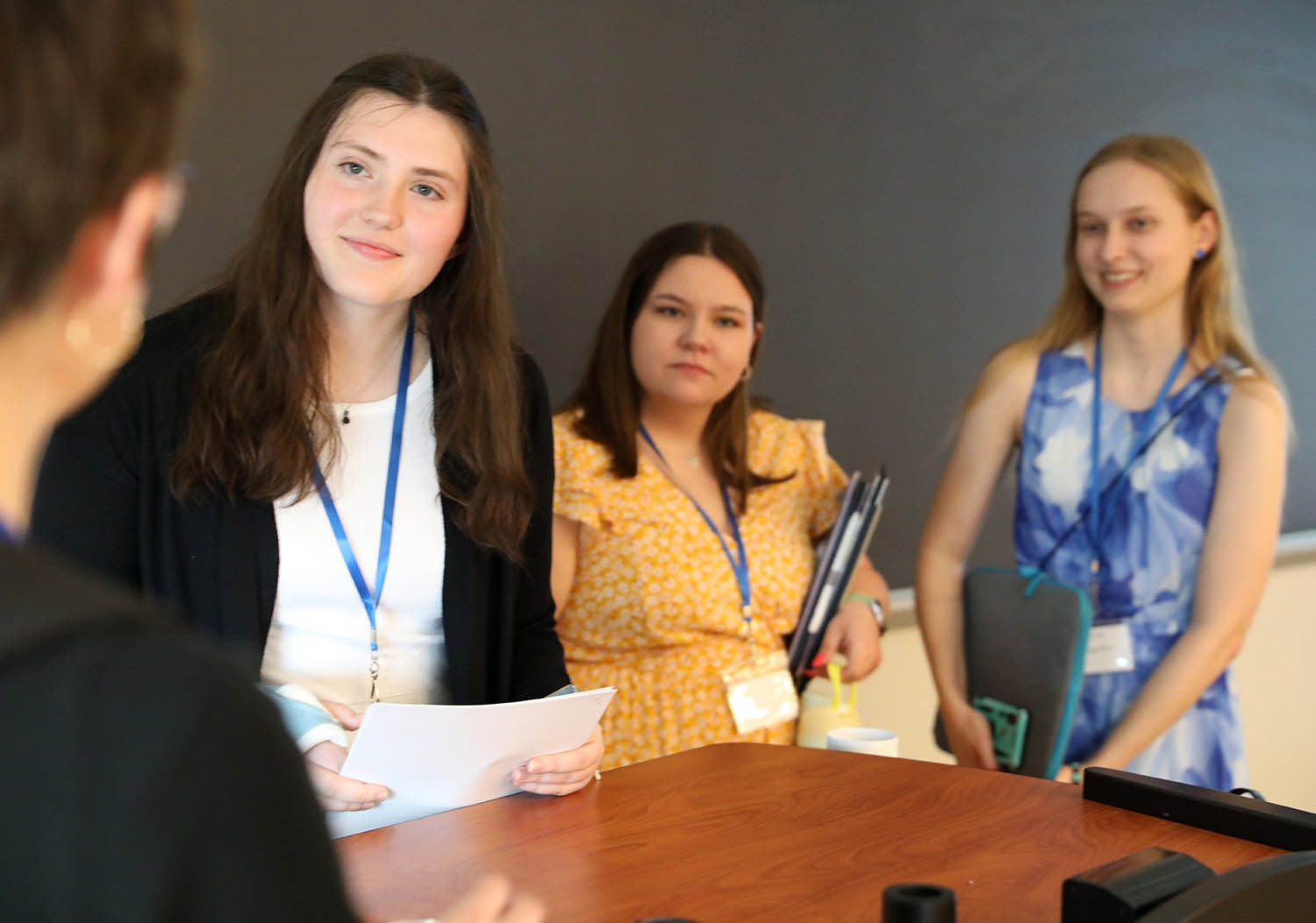 Three young women wearing lanyards, two smiling, one holding papers, another a folder, and a third a laptop sleeve, at a table in a room with a dark wall.