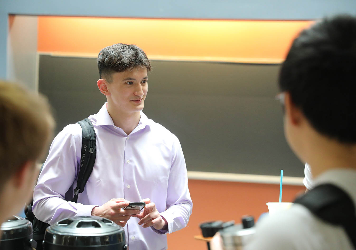 A young man in a light purple shirt and backpack, holding a smartphone, looks right with a slight smile in a classroom.