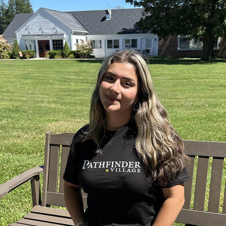 Portrait of Madison Tobon in a black 'Pathfinder Village' t-shirt sits on a bench outdoors, smiling, with a white building and green lawn behind her.