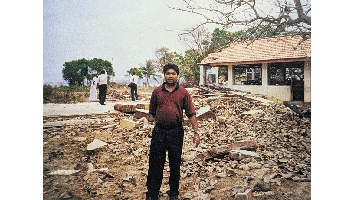 A man in a red shirt stands in front of a large pile of rubble and a partially destroyed building, with other people in the background.