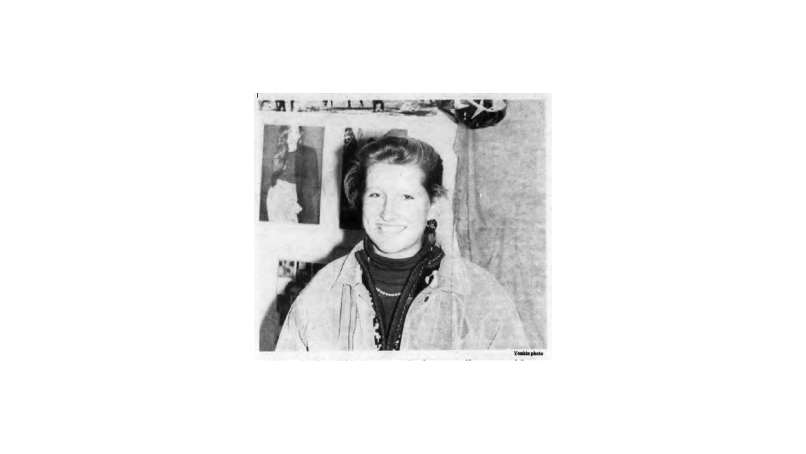 Black and white photo of a smiling young woman with light hair, wearing a jacket, turtleneck, and beaded necklace. Posters and a star are in the background.