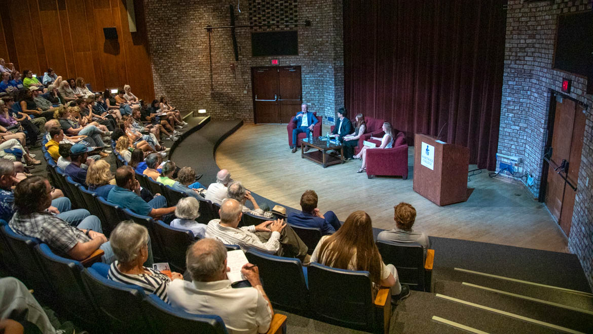 An audience watches a four-person panel discussion with Mike McIntire on a stage in an auditorium.