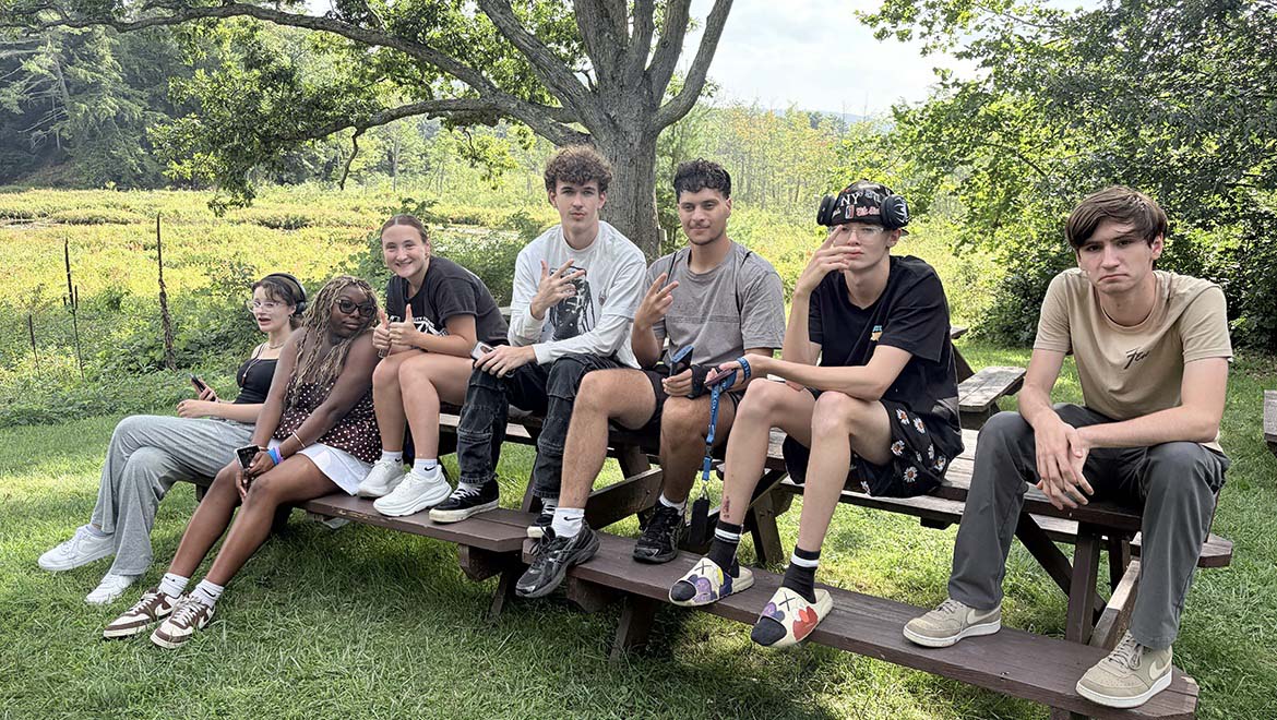 Seven young people sitting on a wooden picnic bench outdoors, surrounded by green trees and plants.