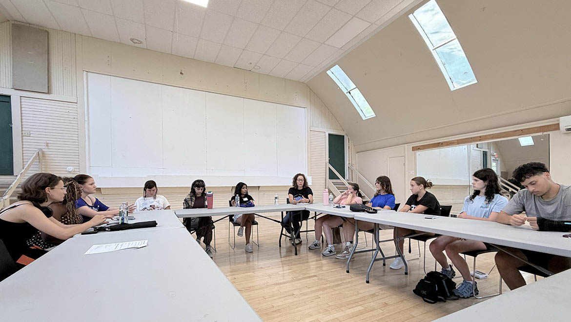 Twelve young adults seated around a U-shaped table in a room with skylights.