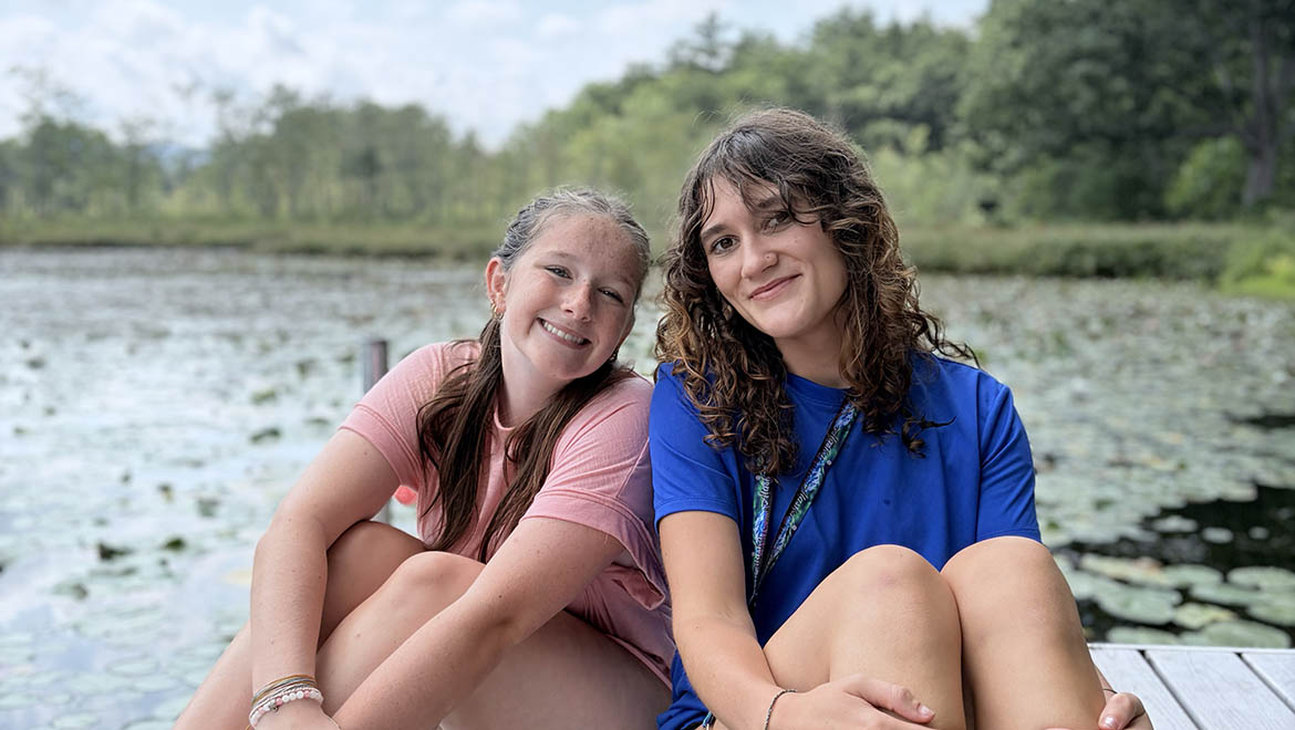 Two young women smiling on a wooden dock overlooking a lake covered in lily pads, with trees in the background.
