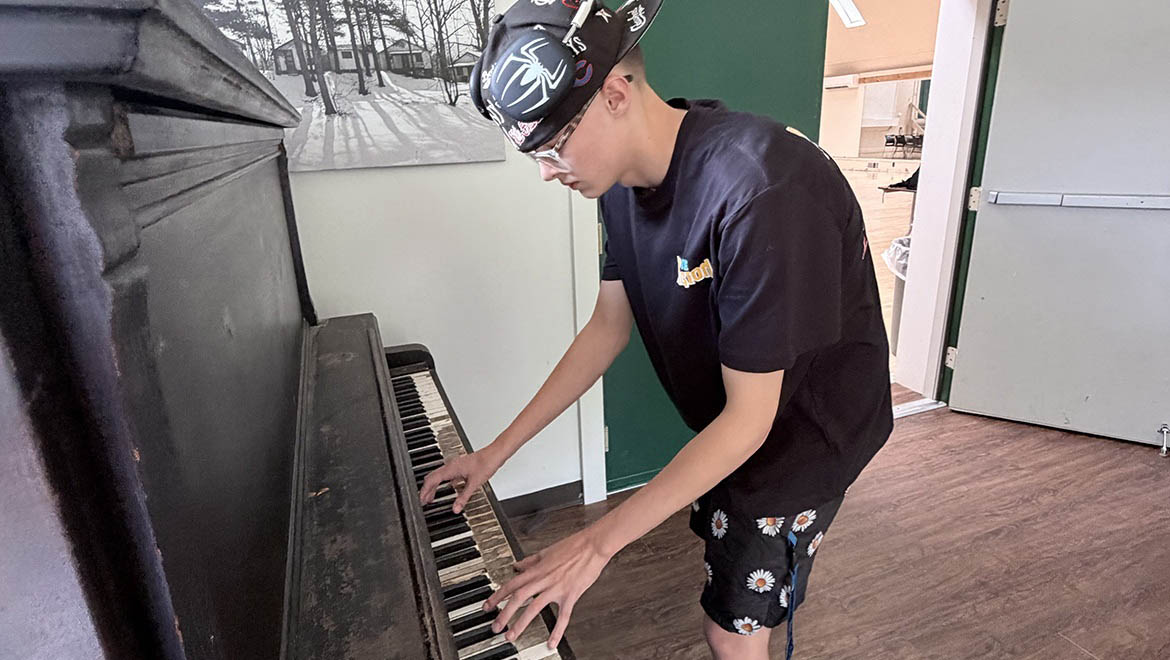 A young person in a Spider-Man hat and daisy shorts plays an old, dark upright piano with worn keys.