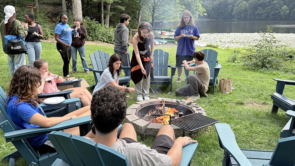 A diverse group of young people gathered around a campfire, roasting marshmallows, with a lake and trees in the background.